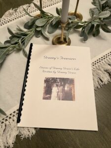 A book titled "Granny's Treasures: Stories of Granny Grace's Life Written by Granny Grace" rests on a wooden table, surrounded by a decorative eucalyptus garland and two