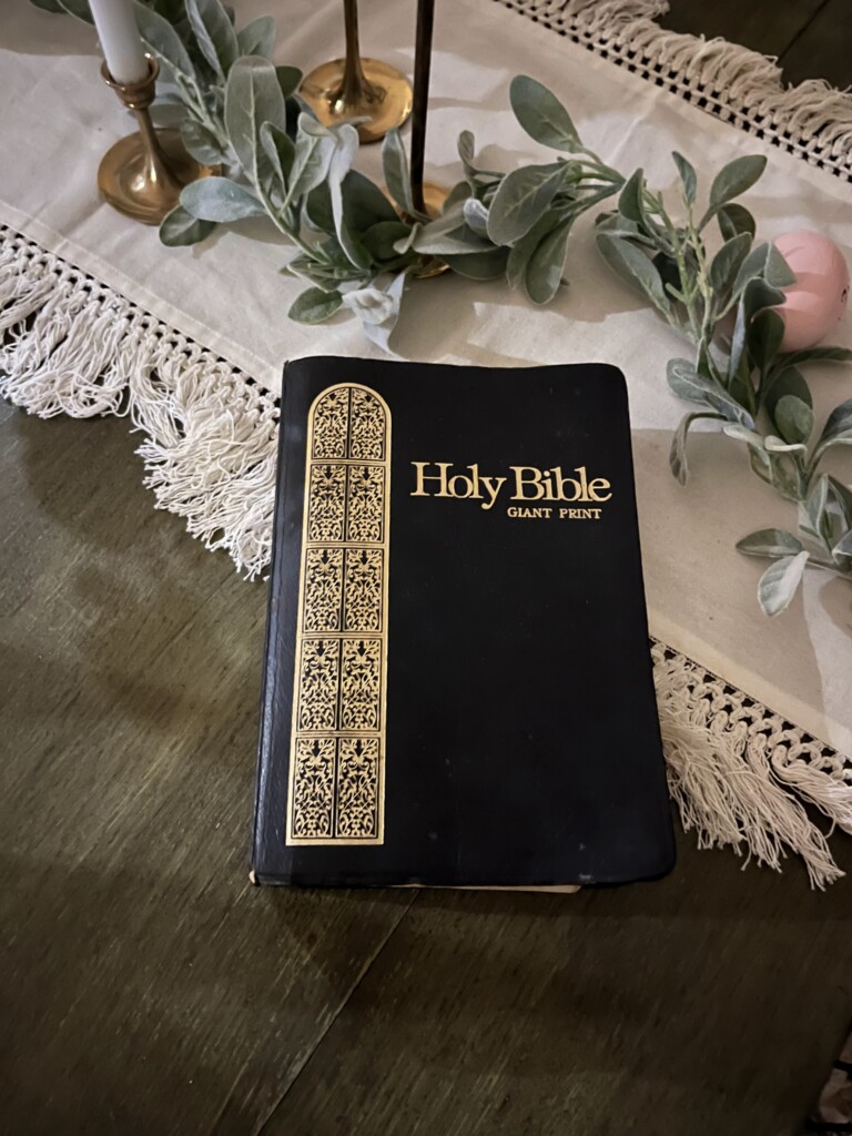 A closed holy Bible with "giant print" on the cover, placed on a wooden table alongside a decorative lace table runner, brass candlesticks, and a eucalyptus garland – legacy 
