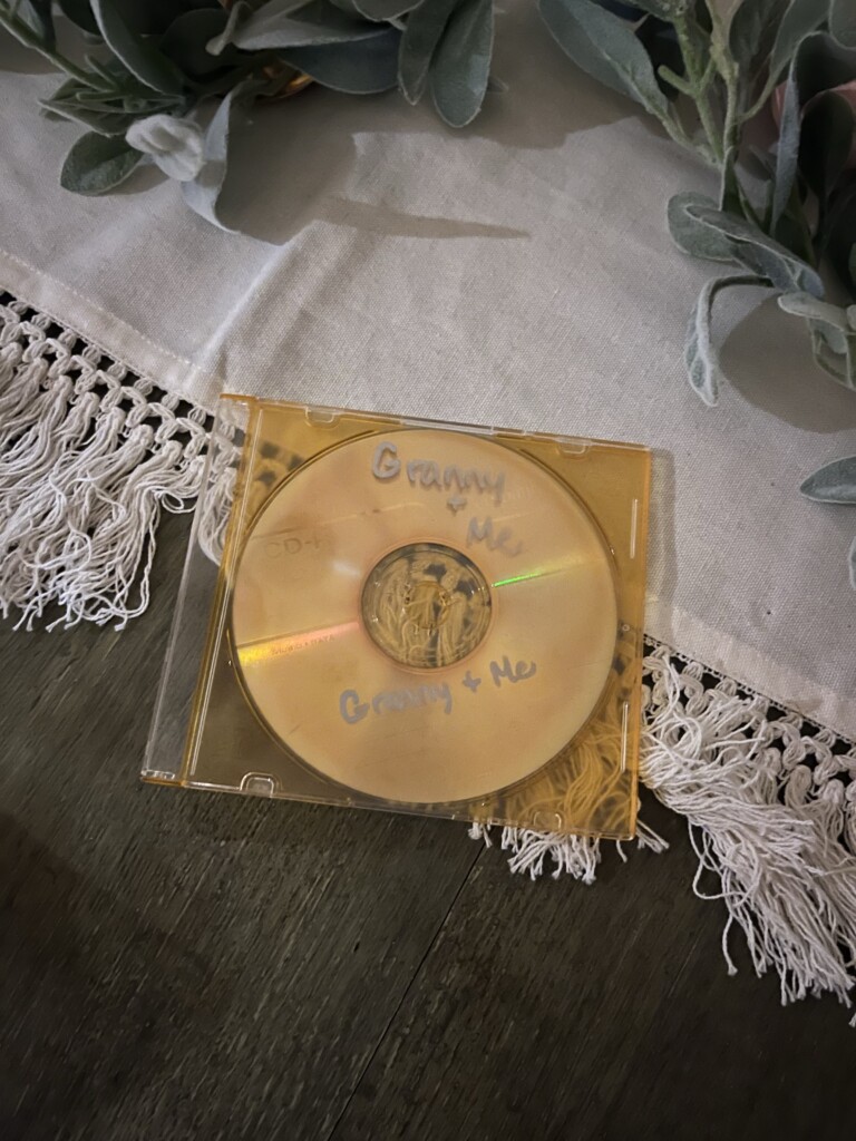 A CD labeled "granny & me" in a clear case, resting on a wooden table with a decorative runner and greenery around it, how to preserve and pass down your family history.