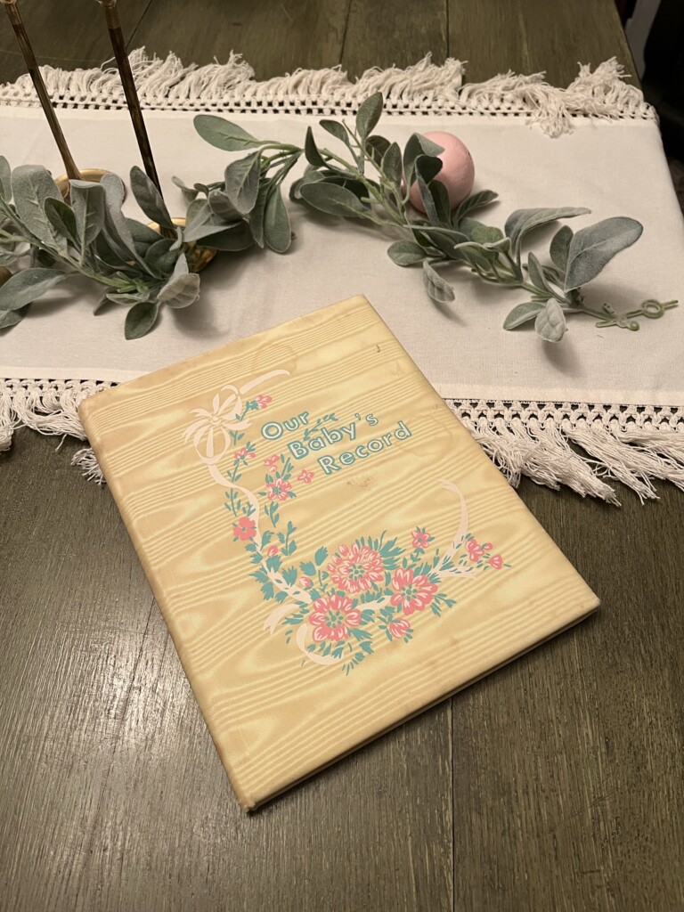 A baby record book with a pastel floral design and the title "Our Baby's Record: How to Preserve and Pass Down Your Family History" on a wooden table, decorated with a simple lace runner