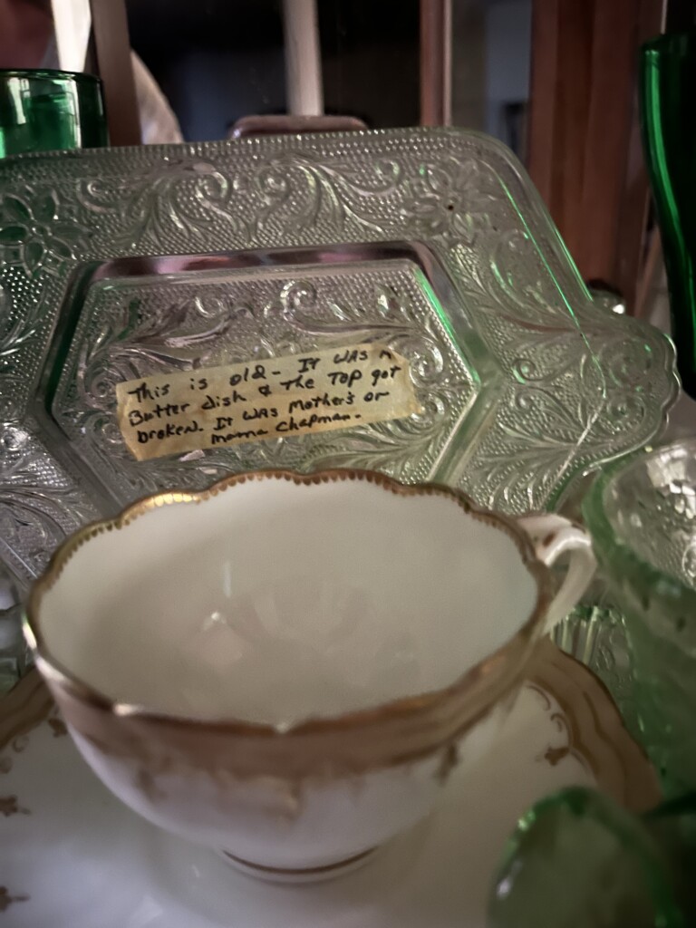 An antique tea cup with a gold rim is displayed in the foreground, with a decorative glass tray and green glassware blurred in the background. A handwritten note on the tray offers historical context related to its