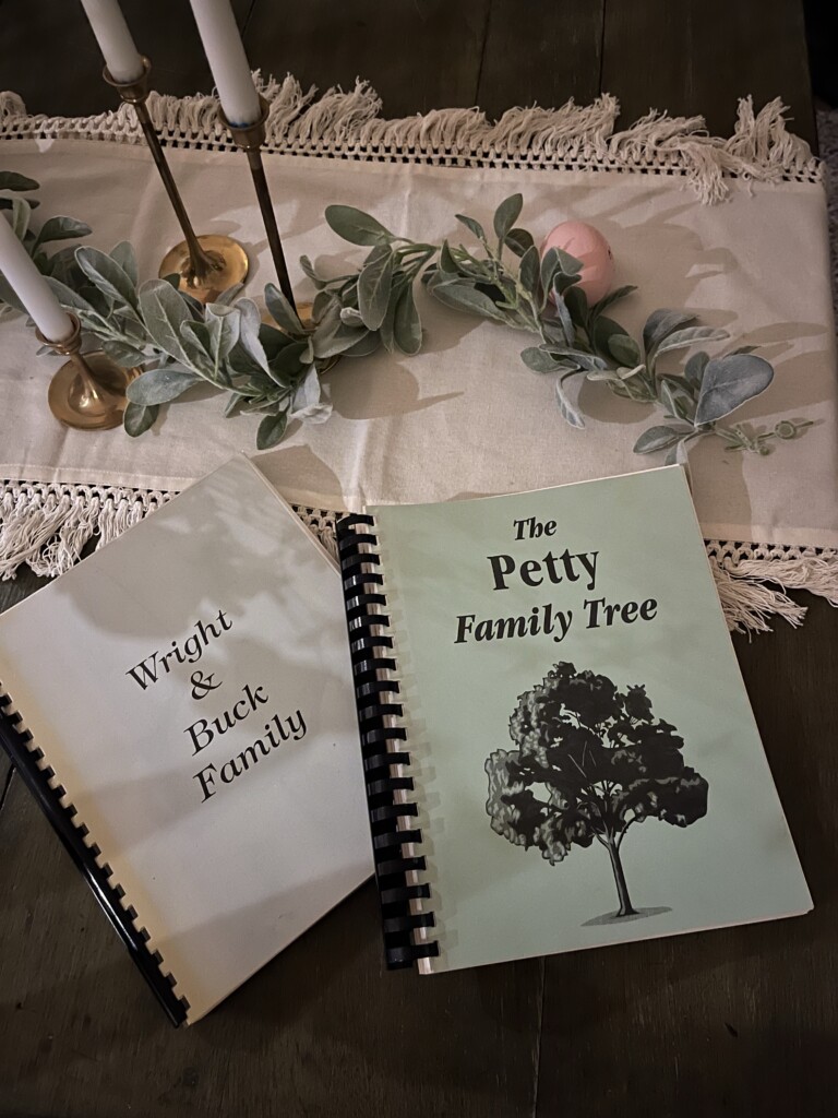 Two family tree books, titled "Wright & Buck Family" and "The Petty Family Tree," on a wooden table with candles and a floral garland, illustrating how to preserve and pass down your