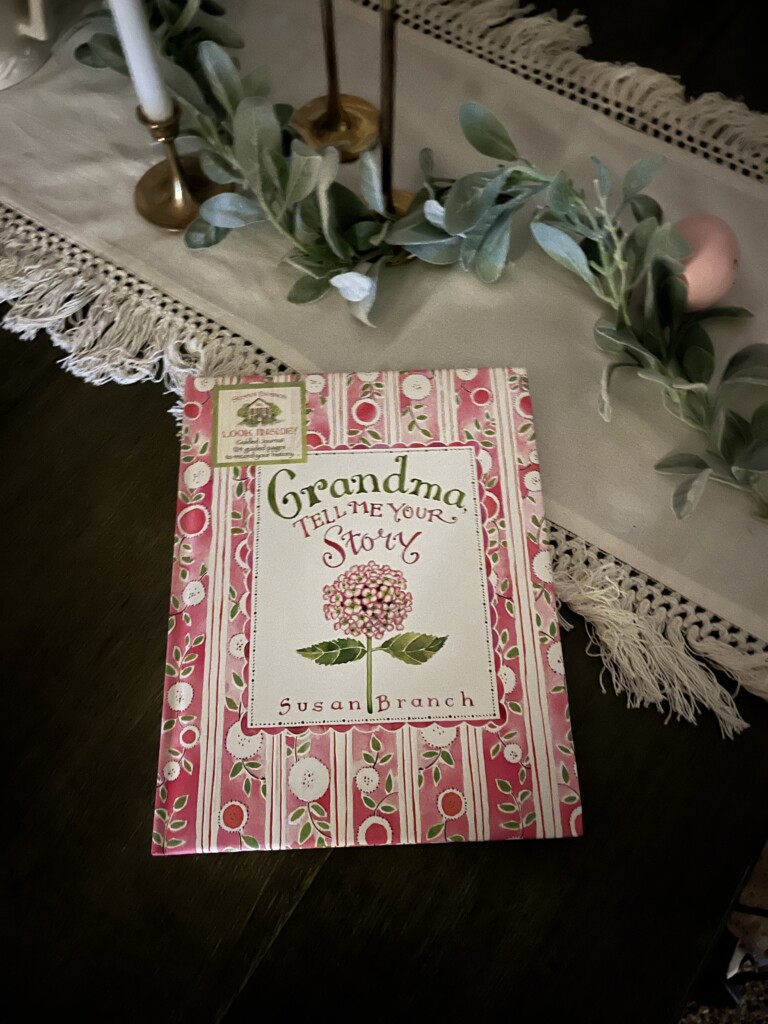 A book titled "Grandma, Tell Me Your Story: How to Preserve and Pass Down Your Family History" by Susan Branch placed on a wooden surface, surrounded by greenery and candles, evoking legacy