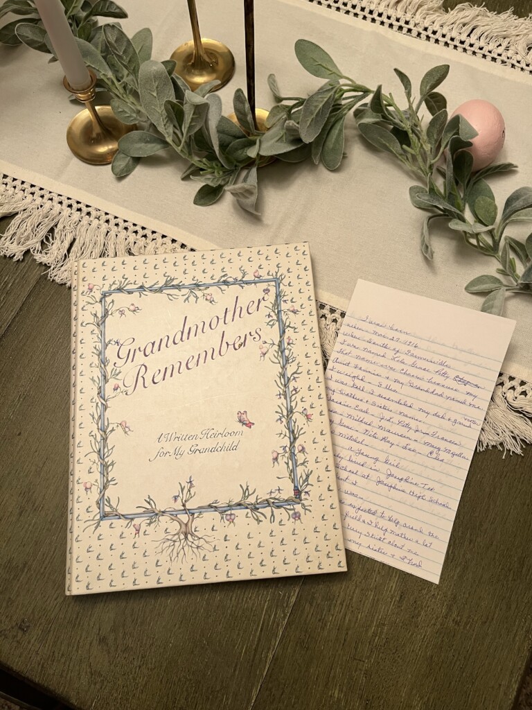 A book titled "Grandmother Remembers: How to Preserve and Pass Down Your Family History"  placed on a wooden surface, surrounded by greenery and candles, evoking legacy