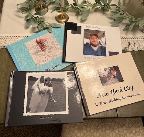 A collection of photo albums and personalized books on a table, showcasing how to preserve and pass down your family history, including wedding, family, and travel themes. Decorative leaves and a candle holder are around them.