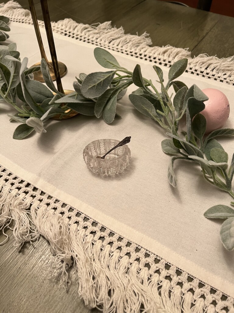 A decorative table setting featuring a glass antique salt dip, a pink egg, and green lambs ear garland on a textured white table runner with fringe edges.
