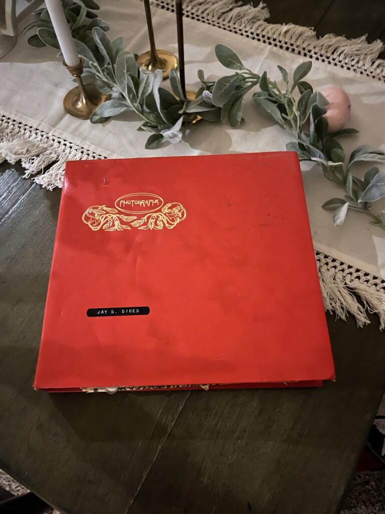 A red scrapbook with golden embossed details, labeled "Jay S. Sikes," rests on a table with a lace tablecloth, flanked by candle holders and greenery, symbolizing legacy