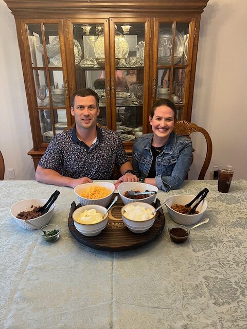 A man and a woman are sitting at a dining table, smiling at the camera. The table is set with bowls containing various ingredients, such as shredded cheese, diced onions, and chopped meat. Behind them is a wooden cabinet with glass panes displaying dishes.