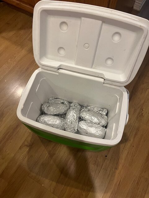 A green cooler with a white lid open, revealing several foil-wrapped items inside, presumably baked potatoes. Perfect for setting up your own baked potato bar! The cooler is placed on a light wooden floor.
