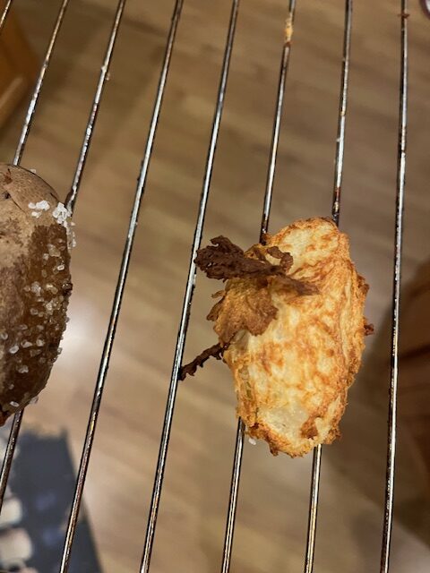 A close-up of what appears to be a baked or fried food item, such as a potato croquette or hash brown. It rests on a metal cooling rack with a wooden floor beneath. Another food item, partially visible on the left, seems to be sprinkled with salt.