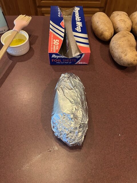 A potato wrapped in aluminum foil is placed on a kitchen counter. In the background, there are a box of Reynolds Wrap foil sheets, a small bowl with a brush in it, and several unwrapped potatoes ready for the Baked Potato Bar.