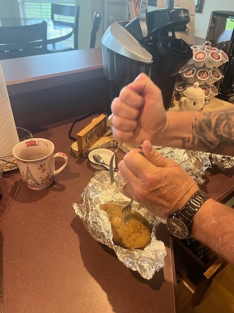 A person with tattooed arms and a watch is seen squeezing a lemon over a baked potato from the baked potato bar, wrapped in foil on a counter. Nearby, there is a coffee maker with coffee pods, a ceramic mug, a wooden container, and a paper towel roll.