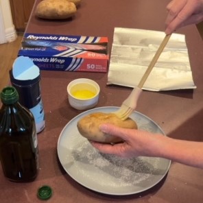 A person uses a brush to apply melted butter to a potato over a plate, preparing for a delicious baked potato bar. Olive oil, a small tub of melted butter, and a salt container are on the counter. Aluminum foil and a box of foil sheets are in the background. Another potato sits on the counter.