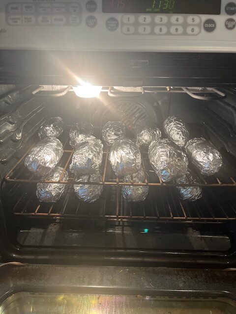 Several potatoes wrapped in aluminum foil are being baked in an oven for a delicious Baked Potato Bar. The oven light is on, and the digital clock on the oven reads 11:30. The foil-wrapped potatoes are placed on two oven racks, ready to be topped with your favorite fixings.