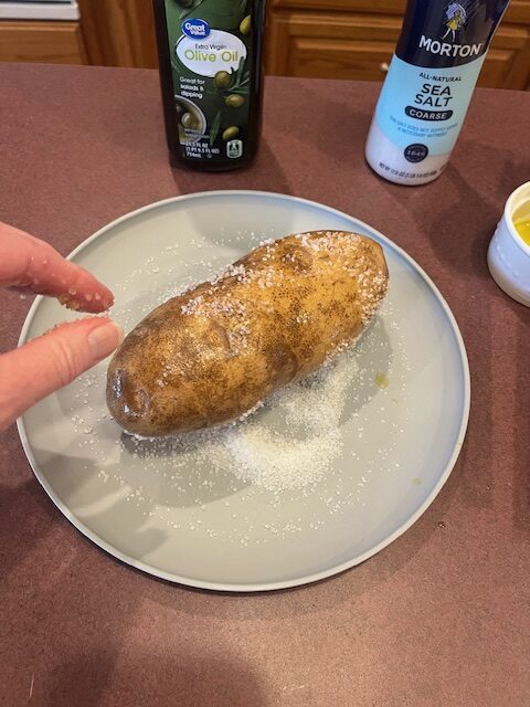 A hand is sprinkling salt onto a potato placed on a grey plate. The potato, destined for a Baked Potato Bar, is already coated with olive oil and some salt. In the background, there are bottles of olive oil and coarse sea salt on a reddish-brown countertop.