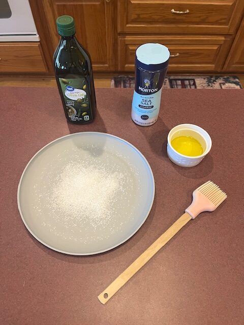 A kitchen countertop showcases cooking ingredients and utensils, ready for a Baked Potato Bar. There is a plate with salt, a small bowl with a beaten egg, a bottle of olive oil, a container of sea salt, and a pastry brush with a light wooden handle and pink bristles.
