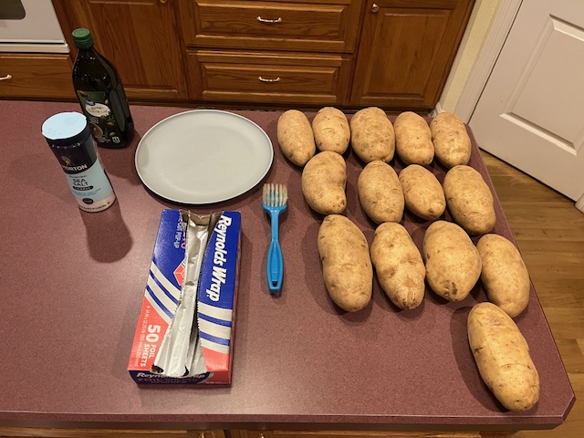 A kitchen counter showcases the ingredients for a delightful Baked Potato Bar: 15 russet potatoes, a bottle of olive oil, a salt container, Reynolds Wrap aluminum foil, a blue brush, and a fork beside a white plate. Wooden cabinets and a door set the background.