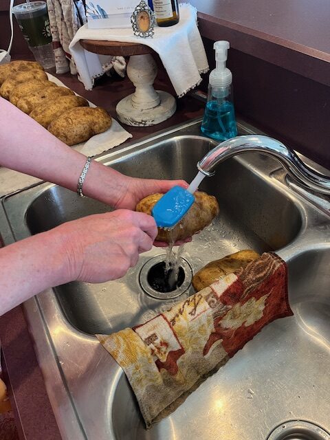 A person wearing a silver bracelet on their left wrist is scrubbing a potato under running water in a kitchen sink using a blue brush. Several other potatoes and a colorful dish towel are visible around the sink area, all being prepped for tonight's Baked Potato Bar.