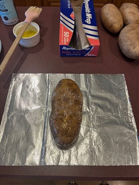 A seasoned baked potato is placed on a sheet of aluminum foil on a countertop. Behind the foil, there is a box of Reynolds Wrap foil sheets, a bowl of melted butter with a brush, and several unseasoned potatoes, perfect for setting up your own Baked Potato Bar.