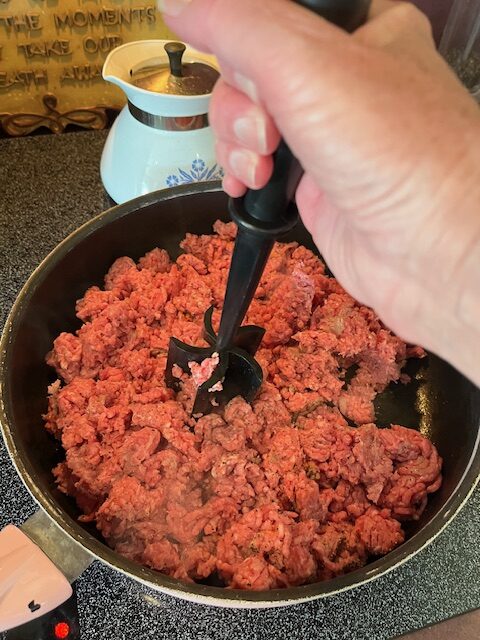 A hand holding a black meat chopper crushes ground beef in a black skillet on a stove, perhaps preparing toppings for an inviting baked potato bar. A white ceramic container with a lid and a metallic pot are visible in the background. The ground beef appears partially cooked.