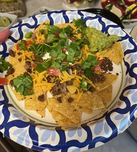 A plate of nachos topped with ground beef, shredded cheese, diced tomatoes, jalapeños, fresh cilantro, sour cream, and a scoop of guacamole. Reminiscent of a Baked Potato Bar, this dish is served on a blue and white paper plate.