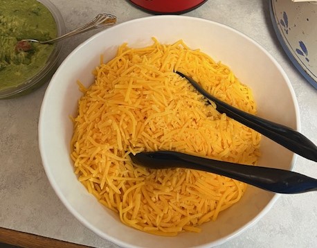 A white bowl filled with shredded cheddar cheese sits on the countertop, perfect for a Baked Potato Bar. Black serving tongs rest atop the cheese, ready for use. Next to it, a spoon peeks out from a separate bowl of guacamole, inviting guests to customize their plates.