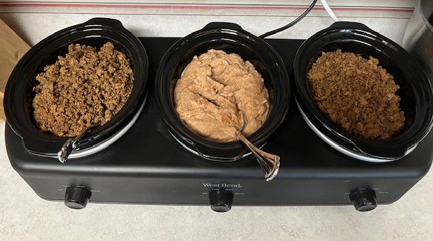 Three black slow cookers on a kitchen counter contain different dishes, perfect for a Baked Potato Bar. The left and right cookers are filled with crumbled ground meat, while the central cooker holds a creamy, light-brown mixture with a spoon in it. All slow cookers rest on a single power base.