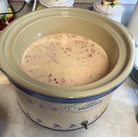 A vintage crockpot, adorned with blue floral patterns, sits on a kitchen counter, ready for a baked potato bar. It is filled with a creamy pink mixture that appears to have chunks of vegetables or meat. Kitchen items, such as canisters and towels, are visible in the background.