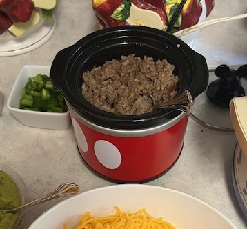 A small red slow cooker with white circles on it holds a cooked mixture, breakfast sausage. Surrounding it are bowls of green bell peppers, shredded cheese, and guacamole, perfect for a Baked Potato Bar set on a light-colored countertop. A colorful ceramic container sits in the background.