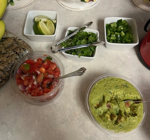 A countertop showcases a variety of taco toppings, including a container of guacamole, a container of salsa, diced green bell peppers, chopped cilantro, and lime wedges. Nearby, much like you’d find at a baked potato bar, tongs are placed for serving the toppings.