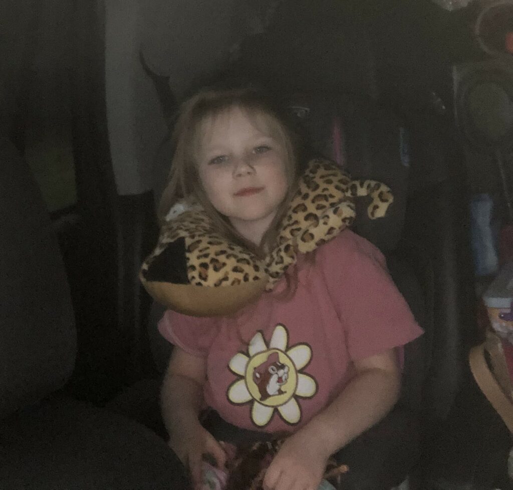 A young girl with blonde hair sits in a car seat wearing a pink shirt with a flower design. She has a stuffed animal neck pillow with a leopard print around her neck and looks at the camera, ready for her beach vacation.