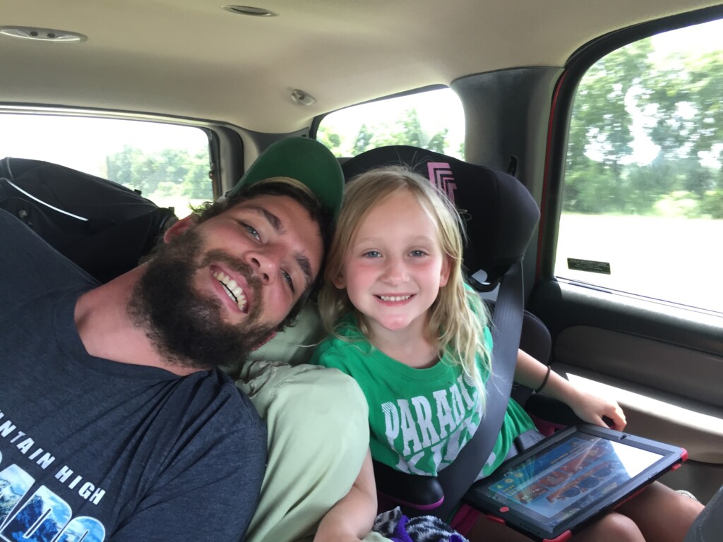 A bearded man in a gray t-shirt leans in close to a smiling young girl in a green shirt who is holding a tablet. They are seated in the backseat of a car, captured in a candid, joyful moment on their beach vacation, with trees visible through the window.