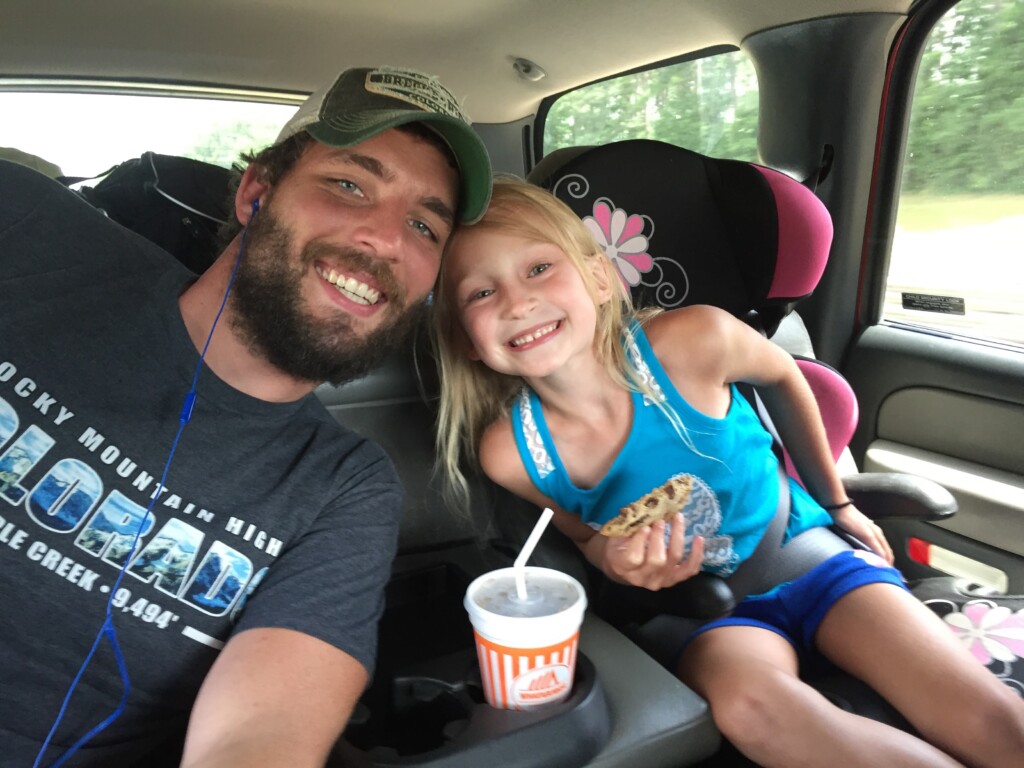 A bearded man wearing a green cap and gray T-shirt poses for a selfie in a car with a young girl who has long blonde hair. The girl is smiling, holding a cookie, and sitting in a car seat. With the drink cup in the cup holder next to her, they look ready for their beach vacation.
