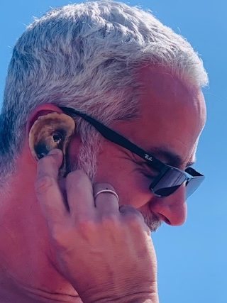 A man with gray hair and sunglasses holds a large seashell up to his ear while smiling under a clear blue sky, enjoying his beach vacation.
