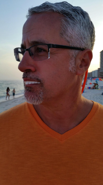 A man with short gray hair and glasses is looking to the side while standing on a beach. He is wearing an orange V-neck shirt. The background, reminiscent of a perfect beach vacation, includes the ocean, beachgoers, and tall buildings.