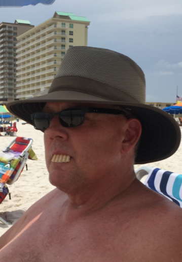 A man wearing a wide-brimmed hat and sunglasses is sitting on a beach chair under a beach umbrella, enjoying his beach vacation. He has novelty fake teeth protruding from his mouth. In the background are colorful beach towels, umbrellas, and a tall building along the sandy beach.