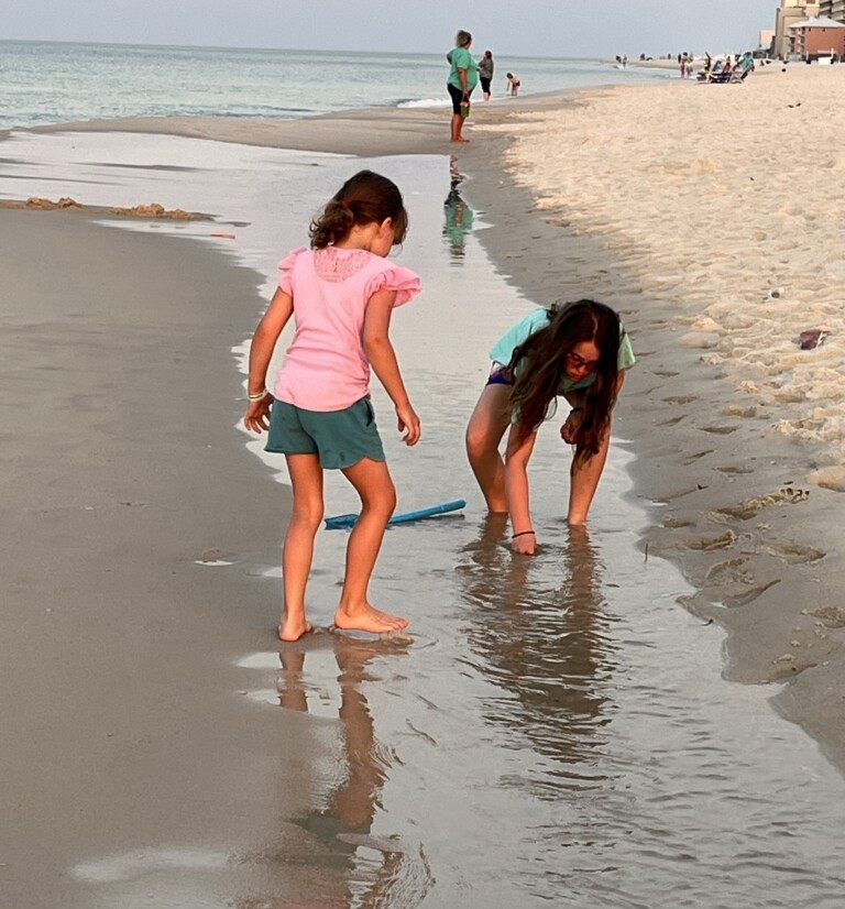 Two young girls enjoy a beach vacation, with one standing in a pink shirt and green shorts, and the other crouching in a green top and purple bottoms, reaching into a shallow water pool. The ocean and other beachgoers are visible in the background.