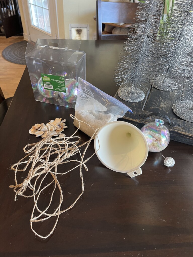 A table with DIY ornament supplies including clear iridescent ornaments, a plastic funnel, a sealed bag of fake snow, twine, decorative flowers, and silver glitter. Silver tabletop Christmas trees and a chair reminiscent of a cozy beach vacation are in the background.