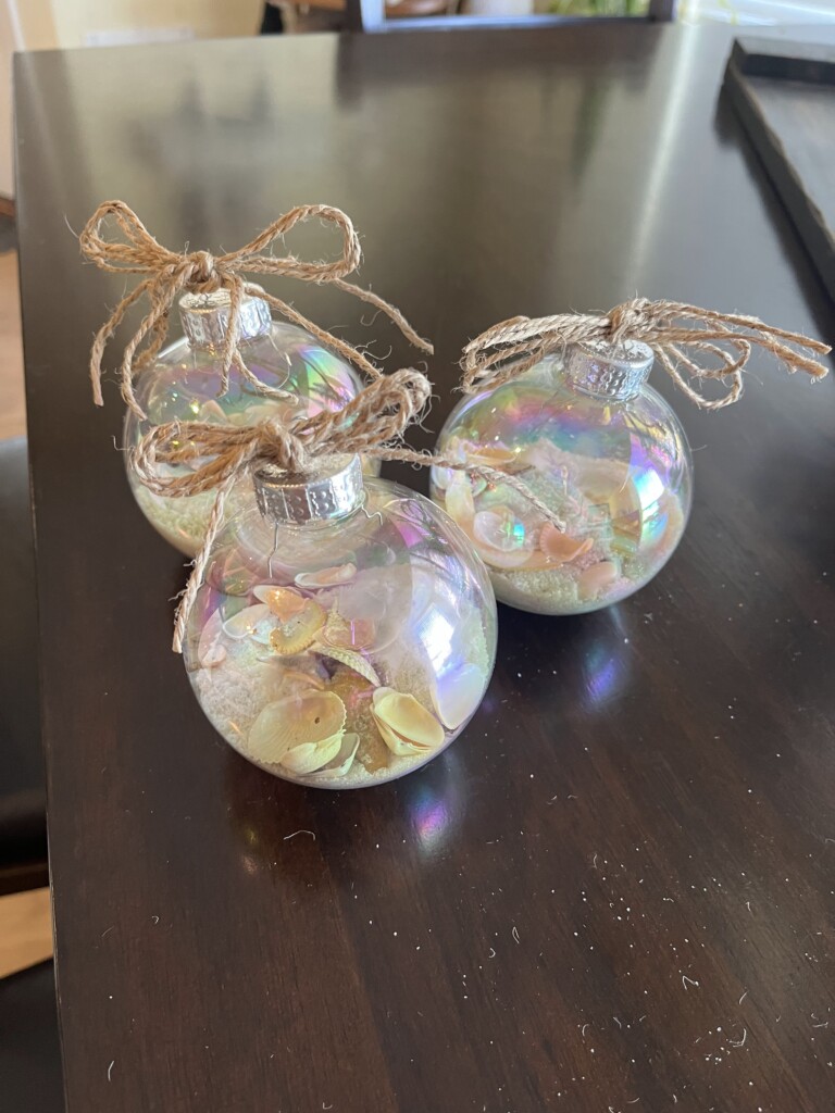 Three clear, iridescent Christmas ornaments sit on a dark wooden table. Each ornament is filled with seashells and sand, reminiscent of a beach vacation, and is topped with a silver cap and adorned with a rustic twine bow.
