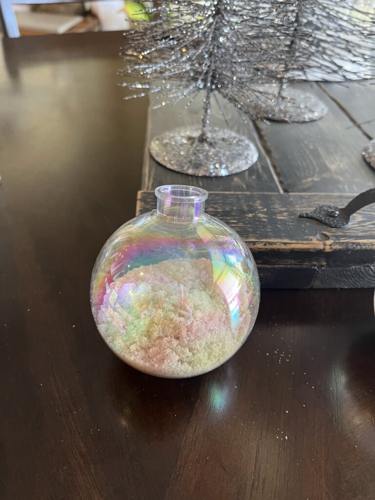 A small, iridescent glass ornament filled with pastel-colored sand sits on a dark wooden table, reminiscent of a beach vacation. In the background, sparkly silver decorative trees add a festive touch.