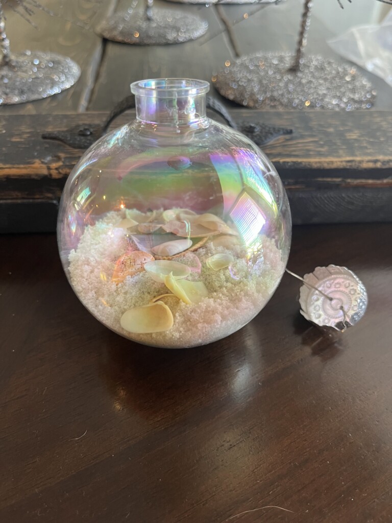 A transparent, iridescent glass ornament sits on a wooden surface. The ornament is filled with small seashells and a layer of white sand-like material, reminiscent of a beach vacation. A delicate, decorative silver tag is attached to the ornament with the word "goodness" engraved on it.