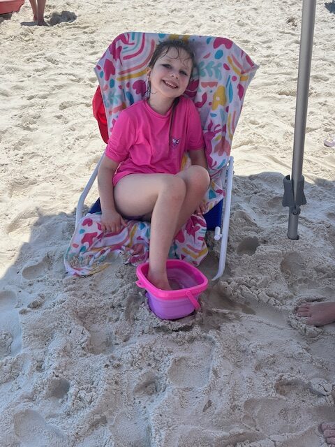 A smiling young girl sits in a beach chair covered with a colorful towel, enjoying her beach vacation. She is wearing a pink shirt and has one foot in a pink plastic bucket filled with sand. The surrounding area is sandy, and part of a beach umbrella and another person's feet are also visible.