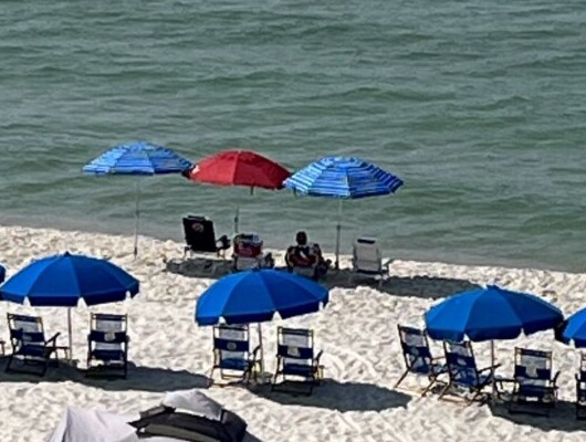 A couple relaxes on beach chairs under two umbrellas, one red and one blue, near the water's edge. Around them, several blue umbrellas and empty beach chairs are scattered on the sandy shore, with the sea visible in the background—an idyllic scene perfect for a beach vacation.