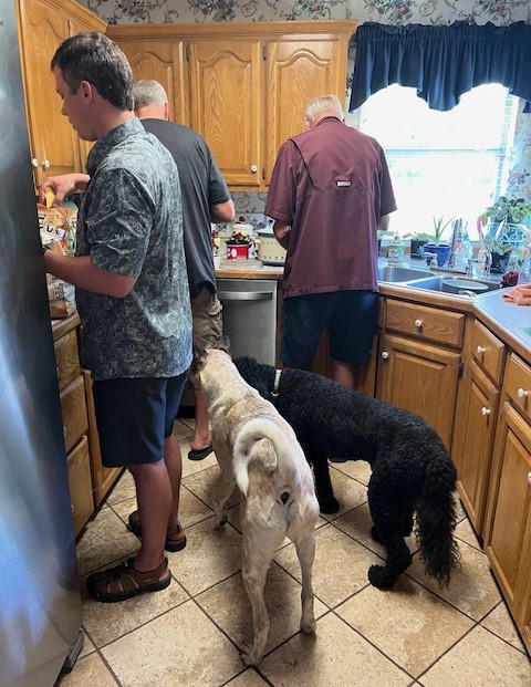 Three men are standing in a kitchen, busy with various tasks at the counter, assembling a DIY Nacho Bar. Two dogs, one white and one black, stand beside them. The kitchen has wooden cabinets and a tiled floor. The men and dogs appear to be intently focused on what they are doing.