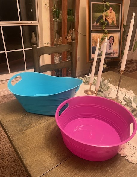 Two plastic tubs, one blue and one pink, are placed on a wooden table next to a centerpiece with greenery and two white taper candles. A chair is positioned behind the table, reminiscent of relaxed evenings after a beach vacation, with framed photos visible in the background.
