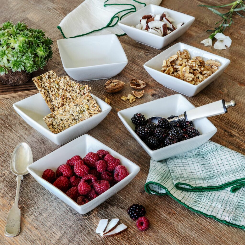 A wooden table with neatly arranged white bowls containing raspberries, blackberries, nuts, granola, and coconut pieces. A spoon lies beside the bowls, and a green and white checkered cloth is nearby. A succulent plant and a white napkin adorn the setting—perfect for a DIY Nacho Bar companion spread.