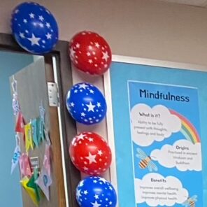 Colorful classroom adorned with red and blue star-patterned balloons on the wall and a "Mindfulness" poster displaying its benefits and tips, giving off the festive feel of a retirement party. International flags hang proudly while a lion plushie sits by a wooden table with a red chair.