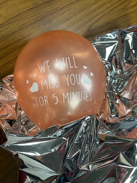 A pink balloon with the words "WE WILL MISS YOU FOR 5 MINUTES" written on it, surrounded by silver, crumpled foil on a wooden surface, perfect for a retirement party setting.