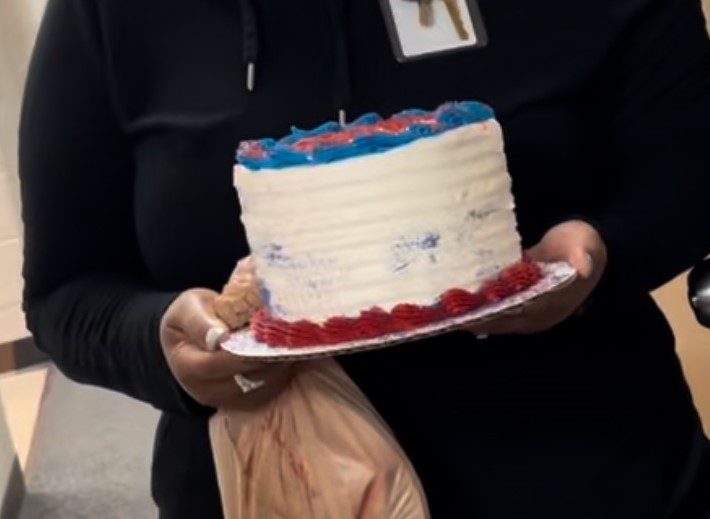 A person wearing a black outfit holds a white cake decorated with red and blue icing at a retirement party. They have light-colored nails and a bag in their left hand. The background is out of focus, showing part of the room they are in. The top of the cake isn't fully visible.