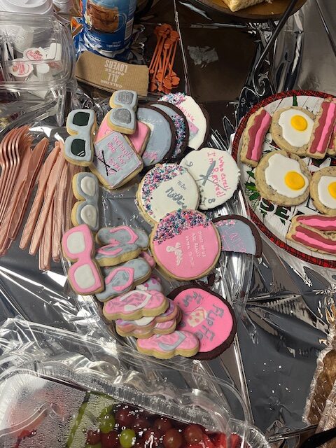 A table with a variety of decorated cookies and silverware: cookies shaped like sunglasses, bottles, and shirts, some with messages like "New Grad," "Buckle Up," and glasses with hands. Plates with bacon and egg cookies are also visible. Pink and silver cutlery is arranged beside them, perfect for a Retirement Party celebration.